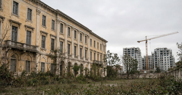 Antigo edifício hospitalar de fachada ocre com jardim abandonado, com prédios de habitação em construção e uma grua ao fundo, num dia nublado