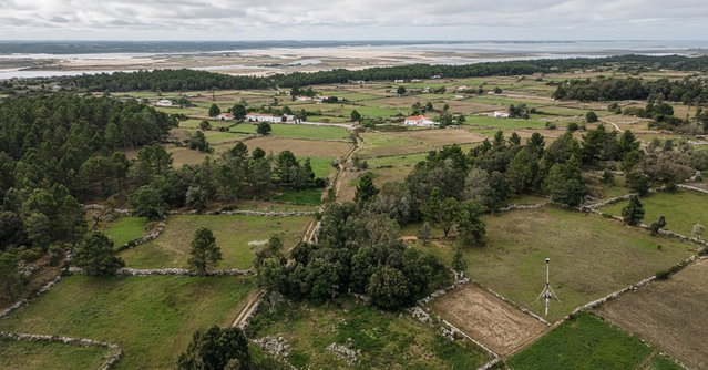 Vista aérea de pequenas parcelas rurais separadas por muros de pedra e caminhos de terra, com pinhal, casas brancas dispersas e uma ria ao fundo, e um recetor GNSS de topografia montado num tripé no canto de um terreno