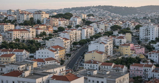 Vista elevada ao fim da tarde sobre bairros residenciais da área metropolitana de Lisboa, com prédios de habitação de várias épocas e uma avenida arborizada