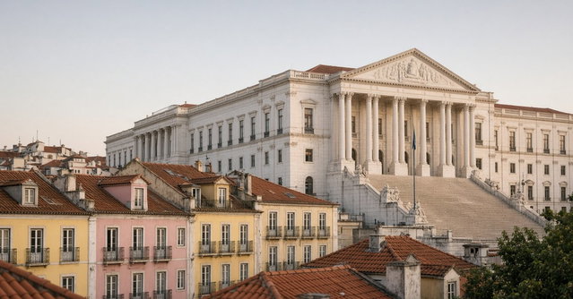 Palácio de São Bento, sede da Assembleia da República, em Lisboa, ao fim da tarde, com telhados de telha e prédios residenciais antigos de fachadas amarelas e cor-de-rosa em primeiro plano