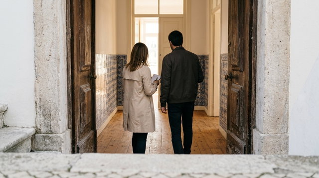 Casal de costas a entrar num apartamento antigo com corredor de azulejos azuis e chão de madeira, durante a visita a uma casa