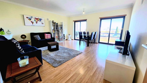 Sala ampla com chão de madeira, sofá azul e sala de jantar junto à varanda, apartamento T3 em Portimão